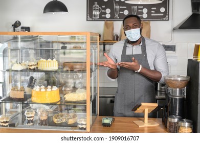 An African American male waiter at the counter wearing medical mask points to fresh pastry and cakes on the showcase. A protective measures for cafe staff during lockdown, pandemic, epidemic
