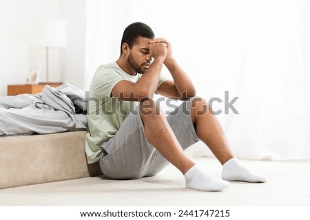 African american man appears downhearted or stressed as he sits next to bed, holding his head, conveying a sense of sadness