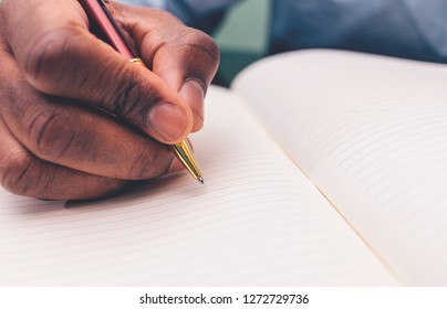 African American Man In Baby Blue Button Shirt Writing In Journal With Red Pen Using Right Hand Room For Print