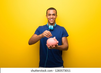 African american man with blue t-shirt on yellow background taking a piggy bank and happy because it is full
