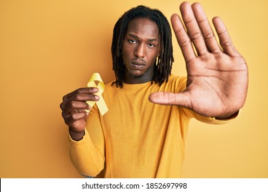 African American Man With Braids Holding Suicide Prevention Yellow Ribbon With Open Hand Doing Stop Sign With Serious And Confident Expression, Defense Gesture 