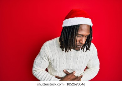 African American Man With Braids Wearing Christmas Hat With Hand On Stomach Because Indigestion, Painful Illness Feeling Unwell. Ache Concept. 