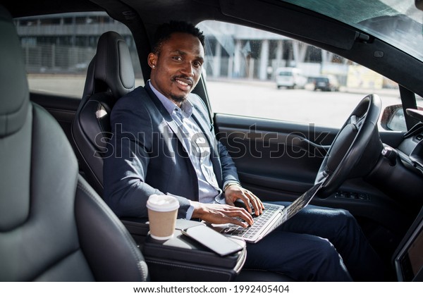 African american man in business suit\
sitting on driver\'s seat of modern electric car and typing in\
wireless laptop. Busy person with portable\
computer.