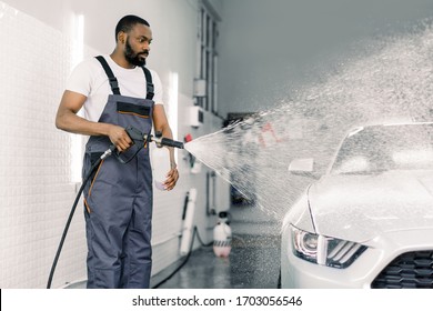 African American man, car wash worker is spraying cleaning foam to a modern white luxury car holding a high pressure washer. Modern car and foam washing, detailing wash