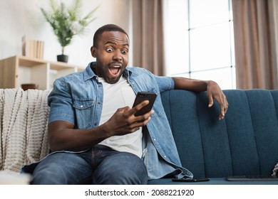 African American Man In Casual Wear Looking On Mobile Screen With Amazed Facial Expression. Happy Young Guy Sitting On Couch Amd Reading Good News On Mobile.