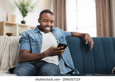 African American Man In Casual Wear Looking On Mobile Screen With Amazed Facial Expression. Happy Young Guy Sitting On Couch Amd Reading Good News On Mobile.