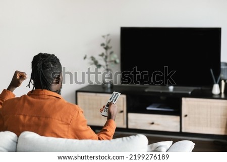 African american man cheerfing for favourite team, watching sports game on TV, celebrating victory, sitting on sofa and looking at television set with empty screen