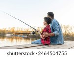 African American man and child sitting on a wooden pier holding fishing rods on the river, dad teaching his son to fish in the lake, family resting and relaxing on the weekend