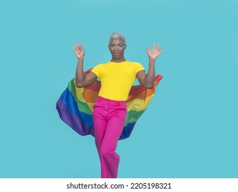 African American Man In Colorful Clothes With Makeup Looking At Camera And Dropping Rainbow Flag Behind Back While Dancing Against Turquoise Background