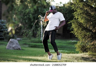 African American Man Dancing Outside In Nature Park. Dancer Wearing White T-shirt, Black Trousers And Baseball Cap