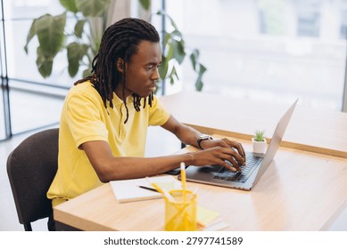 African American man with dreadlocks coding or typing on a laptop, working remotely or as a freelancer in a bright office