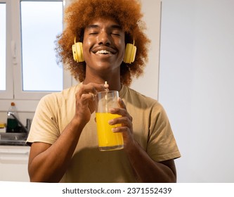 African American man drinking orange juice with a happy face in an apartment while listening to music with headphones - Powered by Shutterstock