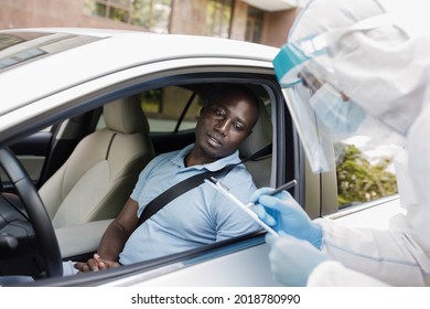 African American Man Driver Sitting Inside White Car, Answering Health Check Up Questions Before Vaccination, Medical Worker In Protective Suit Ticking Off Symptoms On Clipboard Form