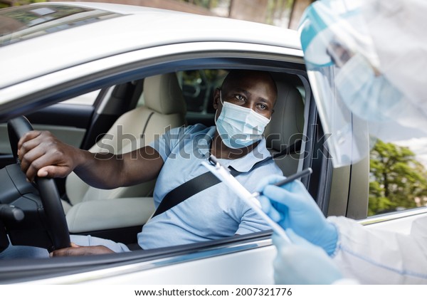 African american man driver wearing protective face\
mask, sitting inside white car, answering health check up\
questions, medical worker in protective suit ticking off symptoms\
on clipboard form