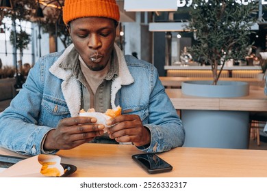 African American man eating burger with fries at table in catering restaurant on mall food court, relaxed atmosphere, urban lifestyle, smartphone on table - Powered by Shutterstock