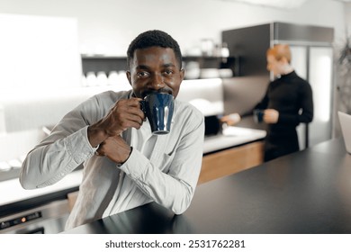 African american man enjoying coffee in modern office kitchen, showcasing relaxed work environment. colleague in background adds to contemporary office scene. - Powered by Shutterstock