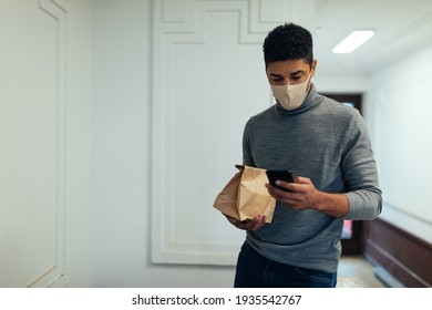 African American Man With Face Mask Using Mobile Phone And Holding Takeout Food.