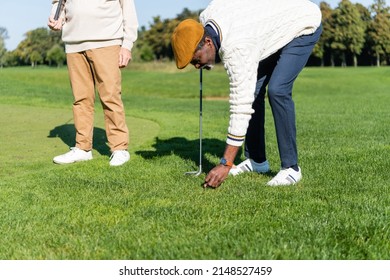 African American Man In Flat Cap Putting Golf Ball On Green Lawn Near Friend