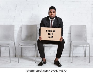 African American Man In Formal Suit Holding LOOKING FOR JOB Sign, Waiting For Work Interview At Office Lobby, Empty Space. Millennial Black Guy Seeking Permanent Employment At Big Company