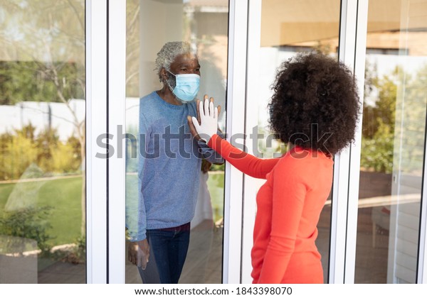 African\
American man greeting his adult daughter through window door,\
wearing face mask. Social distancing and self isolating at home\
during Coronavirus Covid 19 quarantine\
lockdown.