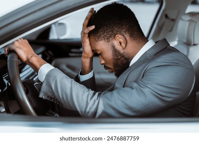 African American man in a grey suit is sitting inside the drivers seat of a car. He has his head in his hands, appearing stressed or frustrated. His hand rests on the steering wheel - Powered by Shutterstock