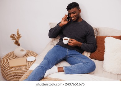 African american man holding coffee and talking on smartphone on bed - Powered by Shutterstock