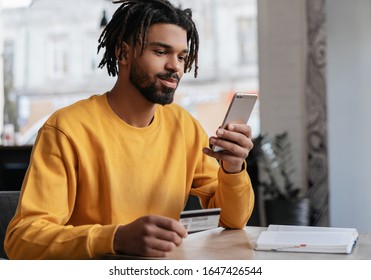 African American man holding credit card, using smartphone for online shopping, mobile banking. Young freelancer receive payment, check balance. Pensive hipster ordering food online, booking tickets - Powered by Shutterstock