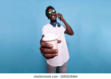 African American Man Holding Cup Of Coffee While Chatting On Phone Call. Black Person With Sunglasses And Headphones Showing Drink To Camera, Talking On Smartphone And Smiling
