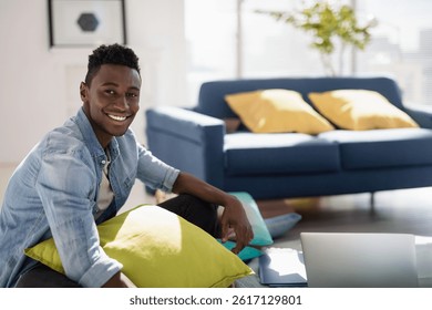 African American man holding green pillow and smiling by laptop in living room, copy space. Interiordesign, homeoffice, remote working, minimalist, sunlit, hardwoodfloor, casual
