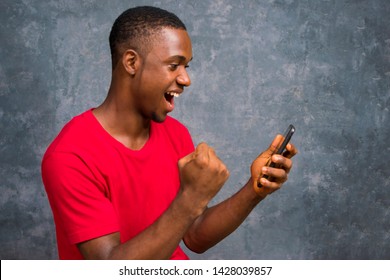 African American Man Holding His Mobile Phone Celebrating His Victory, Success  On Winning A Lottery Ticket. Man Viewing Something Interesting On His Phone