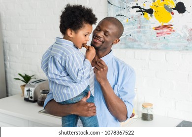 african american man holding little son with cookie in kitchen at home