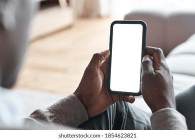 African American Man Holding Smart Phone With Mockup White Blank Display, Empty Screen For App Ads Sitting On Couch At Home. Mobile Applications Technology Concept, Over Shoulder Close Up View.