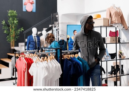 African american man with hood and glasses, trying to steal fashionable merchandise from clothing store. Robber looking around to see if someone watching him while stealing trendy clothes