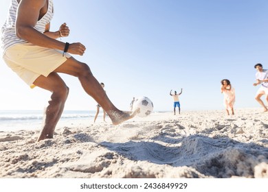 African American man kicks a soccer ball on a sunny beach, wearing yellow shorts. Friends, including a young biracial woman, play in the background, enjoying the leisure time together. - Powered by Shutterstock