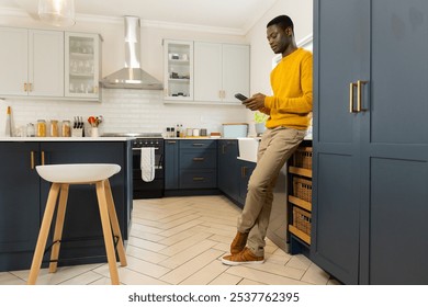 African american man in kitchen using smartphone, wearing yellow sweater, at home. cooking, technology, casual, lifestyle, leisure, relaxation - Powered by Shutterstock