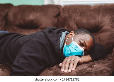 African American Man Laying Down On Couch With Black Sweat Shirt Wearing A Blue Surgical Mask Appearing Sick And Sleepy