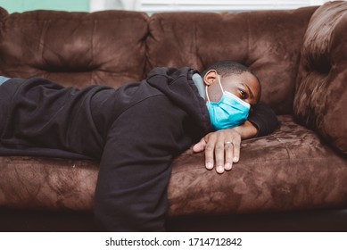 African American Man Laying Down On Couch With Black Sweat Shirt Wearing A Blue Surgical Mask Appearing Sick And Sleepy