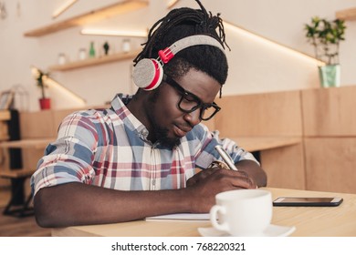 African American Man Listening Music And Writing In Notepad In Coffee Shop