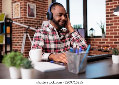 African American Man Listening To Podcast Or Music On Headphones, Using Social Media Online Show On Headset While He Works On Startup Business From Home. Doing Remote Job On Computer.