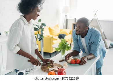 African American Man Looking At Wife Cooking Breakfast In Kitchen At Home