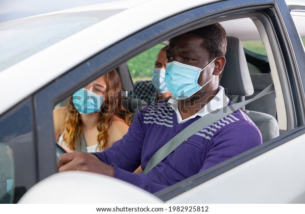African american man in medical\
face mask driving car with friends in passenger seats. Concept of\
new life reality and health protection in coronavirus\
pandemic
