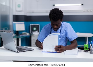 African American Man Nurse Sitting At White Desk In Cabinet While Reading Patient Document Files For Appointment And Checkup. Black Person Analyzing Healthcare Information Papers