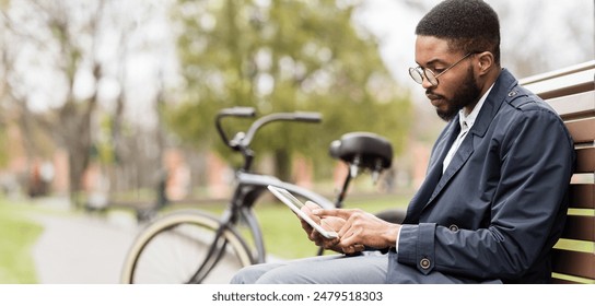 An African American man is on a park bench, working on a digital tablet with a bike nearby. The setting is calm and green, reflecting a mix of business and leisure in nature. - Powered by Shutterstock