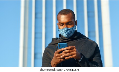 African American Man Out And About In The City Streets During The Day, Wearing A Face Mask Against Air Pollution And Covid19 Coronavirus, Using His Smartphone.