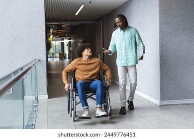 African American man pushing wheelchair of biracial man, both talking in a modern business office. Both wearing casual clothes, African American with dreadlocks, biracial with curly hair, unaltered - Powered by Shutterstock