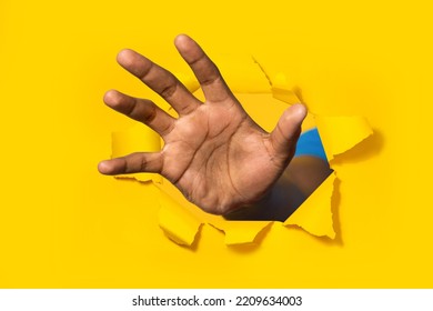 African american man reaching through hole in torn yellow paper, trying to grab something, closeup. Male grasping, trying to touch something, selective focus - Shutterstock ID 2209634003