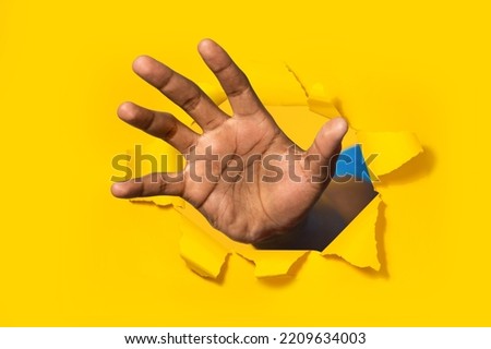 African american man reaching through hole in torn yellow paper, trying to grab something, closeup. Male grasping, trying to touch something, selective focus