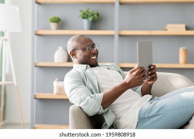 African american man reading e-book on digital tablet or browsing internet, enjoying weekend and resting on armchair at home interior, empty space. Gadget lifestyle, free time concept - Powered by Shutterstock