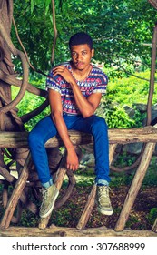 African American Man Relaxing on Park in New York. Wearing a colorful pattern shirt, blue jeans, sneakers, necklace, a young handsome guy is sitting on tree trunk fences, thinking. Instagram effect.
