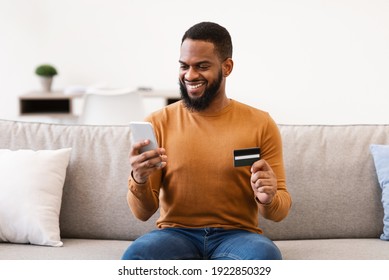 African American Man Shopping Online Using Smartphone And Credit Card Sitting On Couch At Home. E-Commerce And Mobile Banking Application Concept. Selective Focus